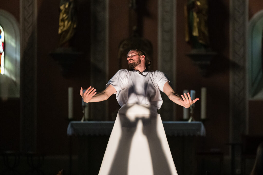 Auf der weißen Kleidung eines Tänzers sind die Silhouetten zweier Hände zu sehen. Der Tänzer steht im Inneren einer Kirche.