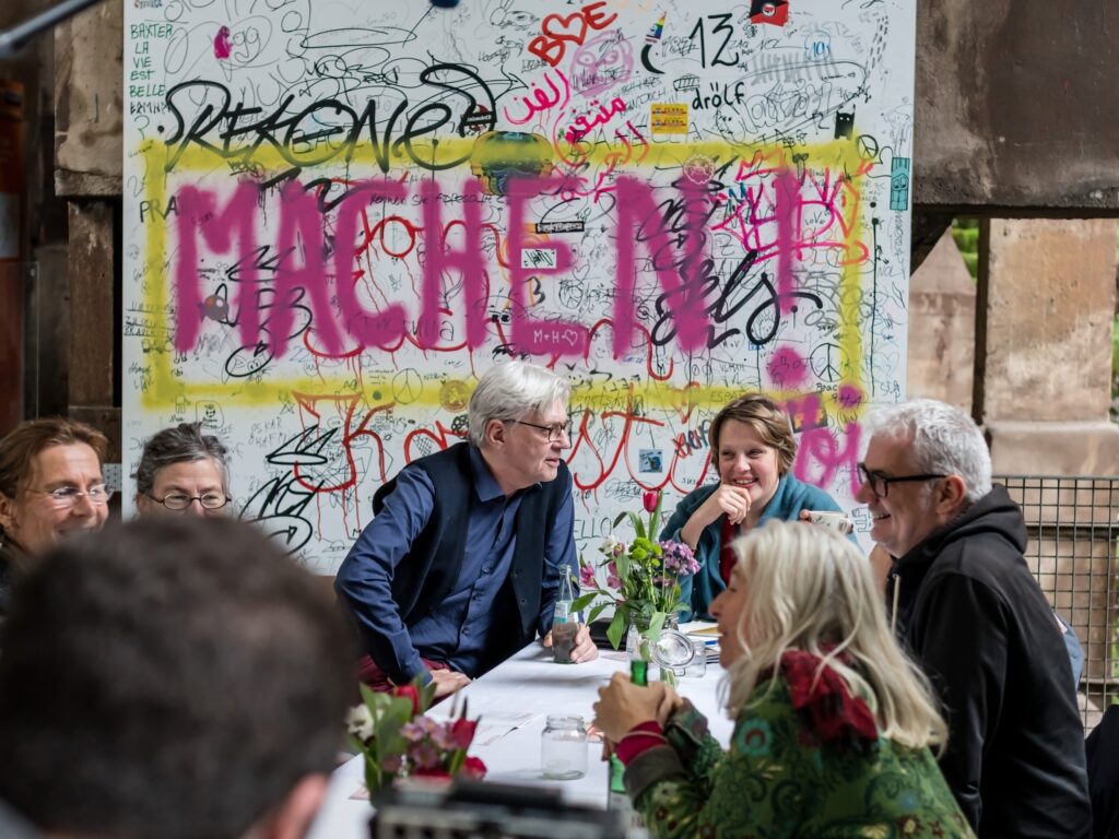 Eine Gruppe von Menschen an einem Tisch diskutiert. Im Hintergrund eine Wand mit dem Shcriftzug: "Machen!"
