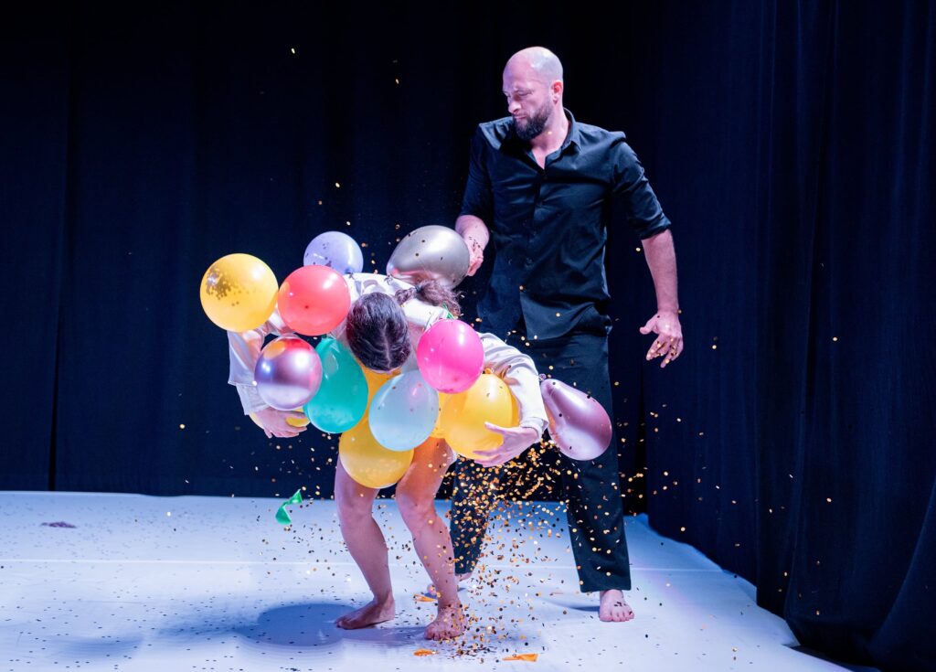 Zwei tanzende Personen auf einer Bühne mit weißem Tanzteppich. Die linke Person hat viele bunte Luftballons im Arm und beugt sich darüber. Konfetti rieselt herunter. Die zweite Person steht dahinter.