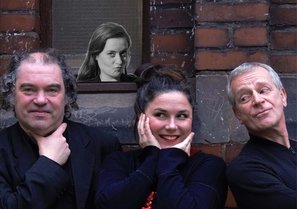 Drei Menschen stehen vor einer Backsteinwand und schauen in die Kamera. Im Hintergrund ist das schwarz-weiß-Foto einer weiteren Person zu sehen.