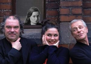 Drei Menschen stehen vor einer Backsteinwand und schauen in die Kamera. Im Hintergrund ist das schwarz-weiß-Foto einer weiteren Person zu sehen.